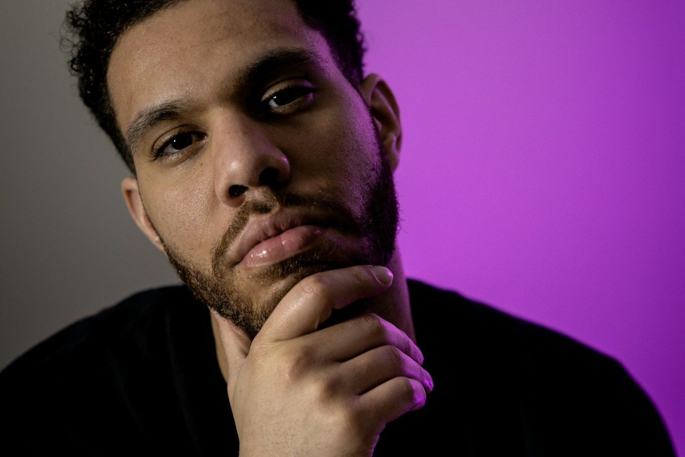 A man against a purple backdrop, looking at the viewer thoughtfully. He has his hand on his chin as though considering something serious